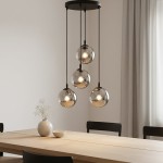 Lampa wisząca Cosmo Premium 4-punktowa w kolorze grafitowym, zawieszona na trzech smukłych linkach. Posiada cztery szklane klosze w kształcie kul, emitujące ciepłe światło, umieszczone nad drewnianym stołem w jasnym wnętrzu.