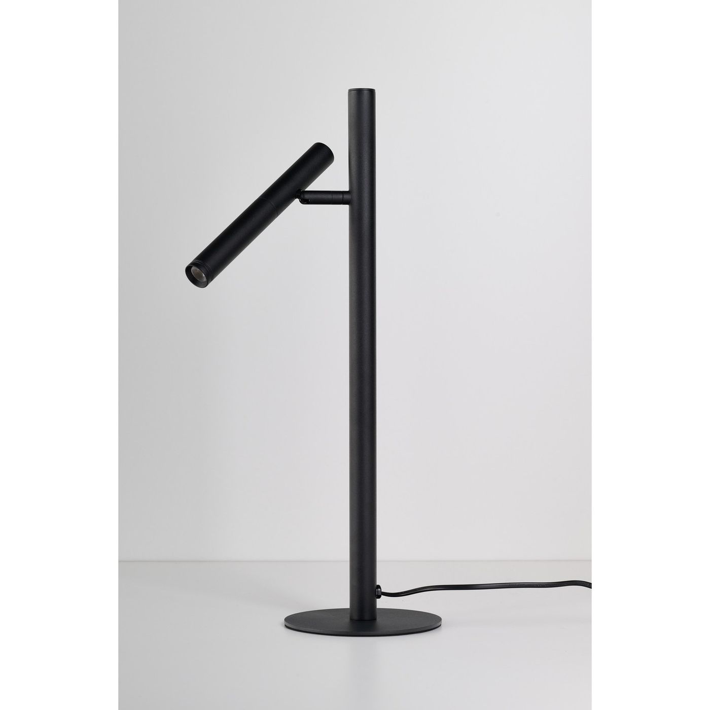 Lampa stołowa LED Trost 4,5W w kolorze czarnym, o minimalistycznym designie. Ujęcie z boku, z pionowym słupkiem i ruchomą głowicą. Stojąca na okrągłej podstawie, z widocznym przewodem zasilającym.