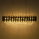 Lampa wisząca Cristal w złotym kolorze, z miejscem na 10 żarówek E14. Posiada dekoracyjne elementy z przezroczystego szkła, wisząca na łańcuchu, układ poziomy, z wyraźnym podziałem na części i teksturą. Tło w odcieniu ciepłego beżu.