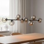 Lampa wisząca Rossi 8-punktowa w kolorze czarnym i grafitowym z szklanymi, przezroczystymi kloszami. Ułożona poziomo, zwisająca z sufitu, z widocznymi żarówkami wewnątrz kloszy. Tło jasne, z drewnianym stołem i zasłonami.