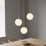 Lampa wisząca Royal 3-punktowa w kolorze białym i złotym z trzema okrągłymi kloszami. Klosze zawieszone na cienkich linkach, w równych odstępach, nad drewnianym stołem w minimalistycznym wnętrzu. Tło w odcieniu szaro-beżowym.