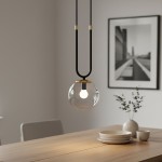 Czarna lampa wisząca Glam 1-punktowa z transparentnym kloszem. Zawieszona na dwóch cienkich przewodach, z metalowym elementem w kolorze złotym. Widoczna żarówka w okrągłym kloszu, otoczenie minimalistyczne, stół z ceramiką w tle.