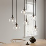 Lampa wisząca Cosmo 6-punktowa z przezroczystymi kulami, zawieszona na cienkich linkach. Kule umieszczone w równych odstępach, z widocznymi żarówkami. Tło jasne, z drewnianym stołem i ceramicznym wazonem.