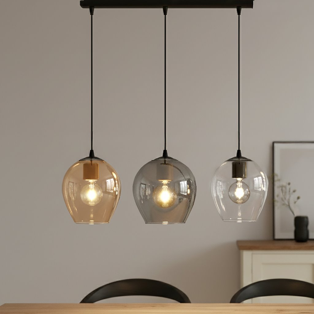Lampa wisząca Istar mix 3-punktowa z trzema szklanymi kloszami w kolorach brązowym, szarym i przezroczystym, zawieszona na czarnych linkach. Klosze mają zaokrąglony kształt, a w środku widoczne żarówki, umieszczona nad drewnianym stołem w jasnym wnętrzu.