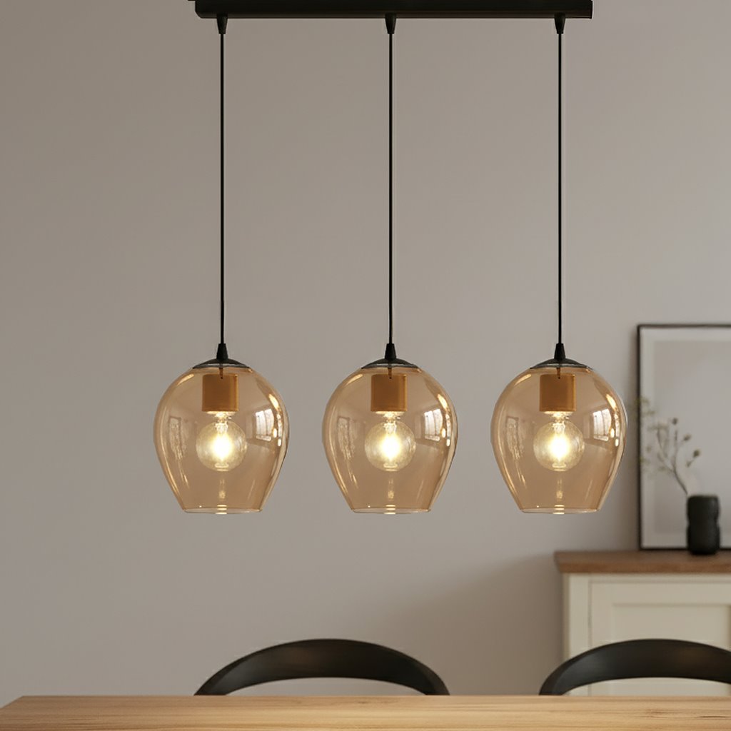 Lampa wisząca Istar 3-punktowa w kolorze miodowym z przezroczystymi kloszami, zawieszona na czarnych linkach. Klosze mają okrągły kształt, a wewnątrz znajdują się żarówki. Produkt umieszczony nad drewnianym stołem w jasnym wnętrzu.