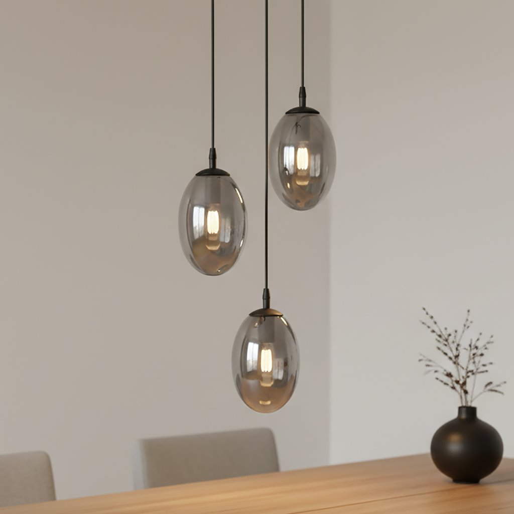 Lampa wisząca Astral 3-punktowa w kolorze czarnym i grafitowym, z trzema owalnymi kloszami w transparentnym szkle, zawieszona na cienkich linkach. Lampa umieszczona nad drewnianym stołem w jasnym wnętrzu.