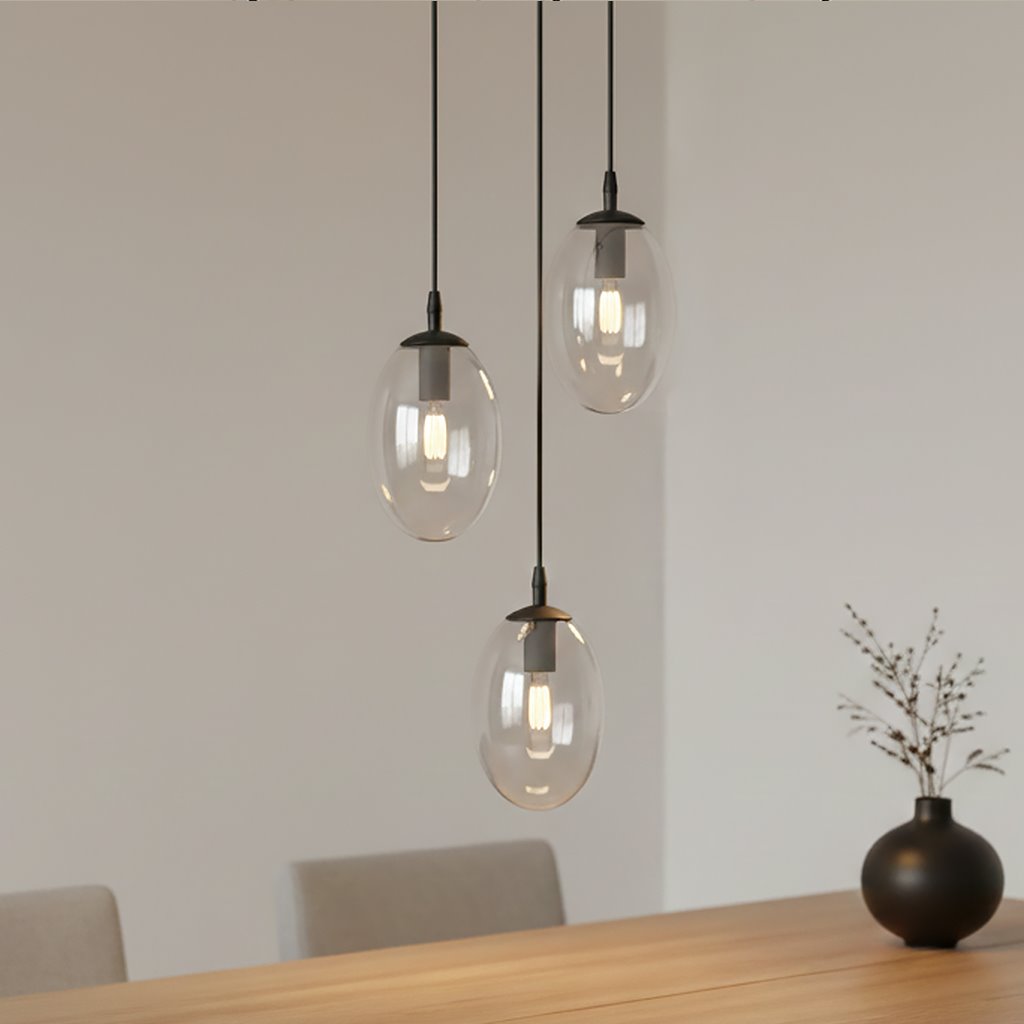 Lampa wisząca Astral 3-punktowa w kolorze czarnym i transparentnym, z trzema szklanymi kulami w kształcie owalu, zawieszona na cienkich kablach. Widoczna żarówka wewnątrz każdej kuli. Tło jasne, minimalistyczne, z drewnianym stołem i wazonem.