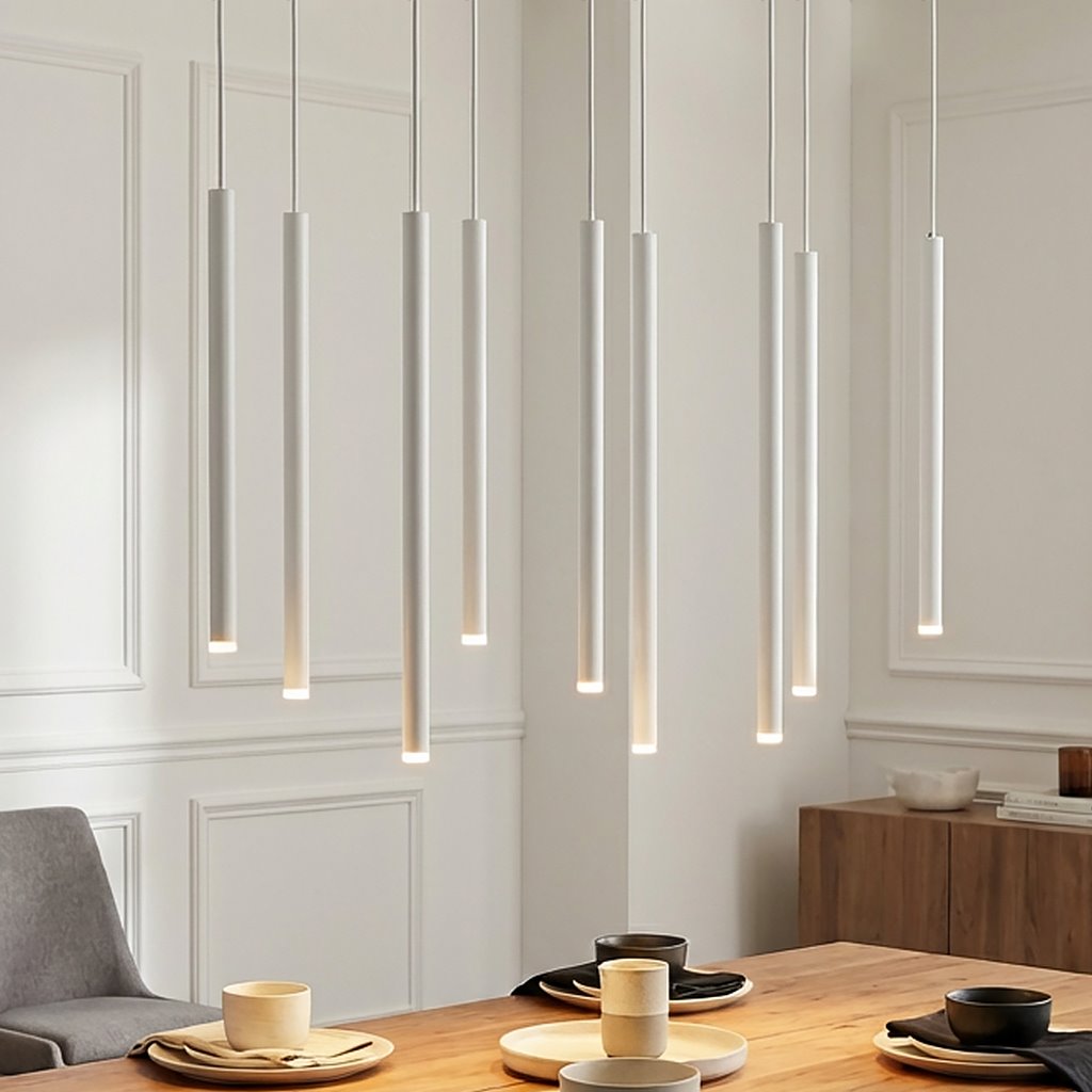 Lampa wisząca Selter 9-punktowa w białym kolorze, składająca się z dziewięciu długich, cylindrycznych elementów świetlnych zawieszonych na cienkich linkach. Produkt umieszczony nad drewnianym stołem, w jasnym wnętrzu z dekoracyjnymi detalami.