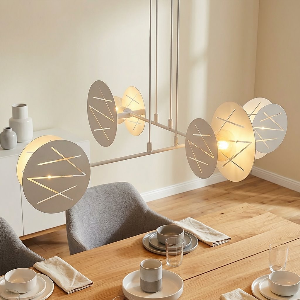 Lampa wisząca Xotic, biała, z czterema okrągłymi kloszami, zawieszona nad drewnianym stołem. Klosze mają geometryczne wzory i emitują miękkie światło. Otoczenie to nowoczesna jadalnia z talerzami, szklankami i serwetkami.