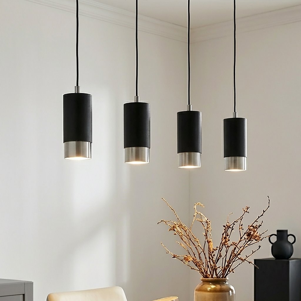 Czarna lampa wisząca Fumiko 4-punktowa z chromowanymi detalami, zawieszona na czarnych przewodach. Lampy mają cylindryczny kształt, a ich dolne części są metaliczne. W tle widać dekoracyjny wazon z gałęziami oraz stół.