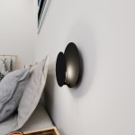 Kinkiet 1-punktowy Idea w kolorze czarnym z okrągłą podstawą, zamontowany na ścianie obok łóżka. Oświetlenie skierowane w dół, z widoczną strukturą materiału. W tle poduszki na łóżku oraz elementy aranżacji.
