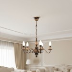 Żyrandol świecznikowy Velita w kolorze patyny, z pięcioma ramionami na żarówki E14. Ujęcie z góry, zawieszony na łańcuchu, w eleganckim wnętrzu z jasnymi zasłonami i sofą w tle. Okrągła podstawa z dekoracyjnymi detalami.