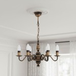 Żyrandol świecznikowy Velita w kolorze patyny, z sześcioma ramionami na żarówki E14. Ozdobny kształt z delikatnymi zdobieniami, zawieszony na łańcuchu. Widoczny na tle jasnych zasłon w eleganckim wnętrzu.