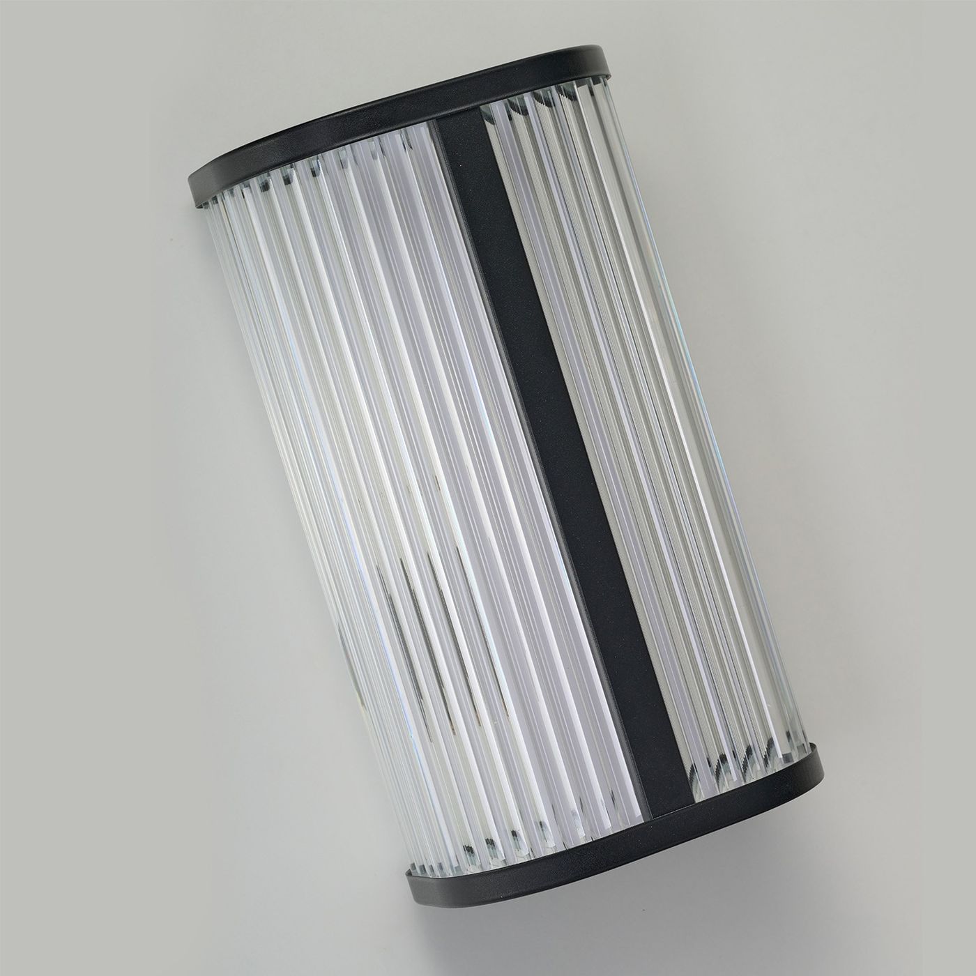 Kinkiet LED Pandora 11W CCT IP44 w kolorze czarnym, o cylindrycznym kształcie, wykonany z przezroczystego materiału, z poziomymi żłobieniami. Ujęcie pod kątem, wiszący na jednolitym tle. Idealny do użytku wewnętrznego i zewnętrznego.