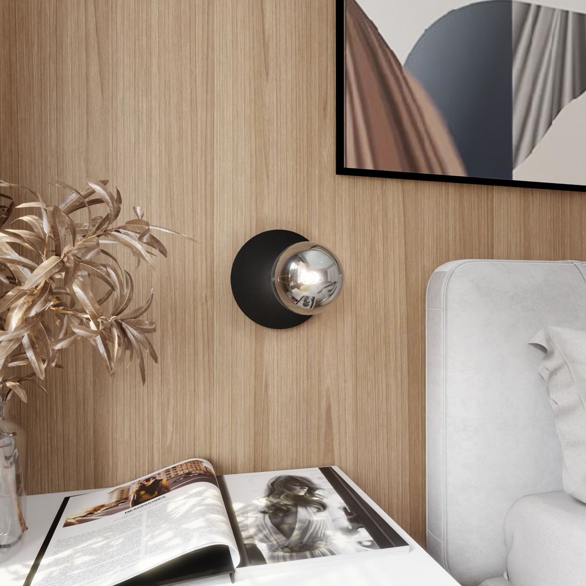Kinkiet Oslo 1-punktowy w kolorze czarnym z grafitowym wykończeniem, z kulistą żarówką. Zamontowany na ścianie, w otoczeniu drewnianej struktury oraz białych mebli. W tle widoczna gazeta na stole oraz roślina dekoracyjna.