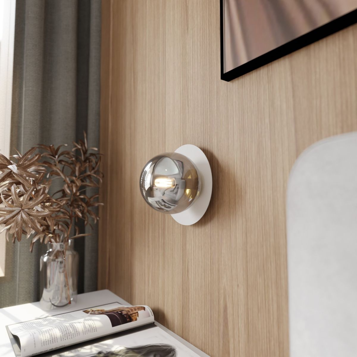 Kinkiet Oslo 1-punktowy w kolorze białym i grafitowym, zamontowany na ścianie, z okrągłą, przezroczystą osłoną lampy. W tle drewniana ściana oraz stół z magazynem i wazonem z dekoracyjnymi gałązkami.
