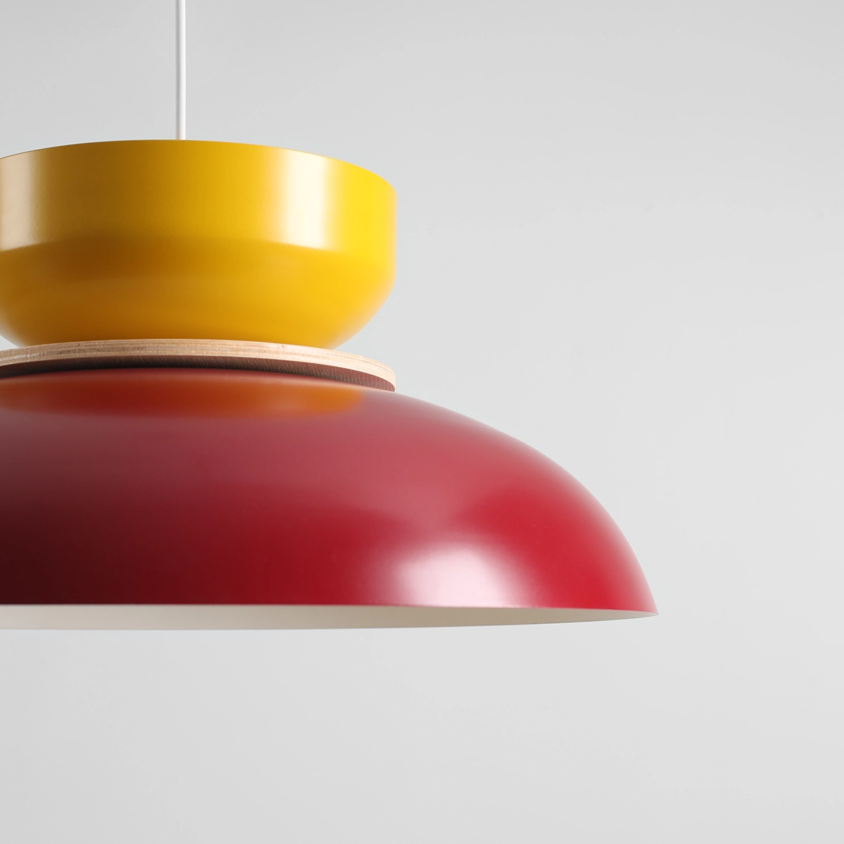 Lampa wisząca Aldo z jedną oprawą E27 w kolorze czerwonym w kształcie półkuli, z żółtą, okrągłą górną częścią. Elementy połączone drewnianym pierścieniem. Tło jasnoszare, lampa zawieszona w powietrzu.