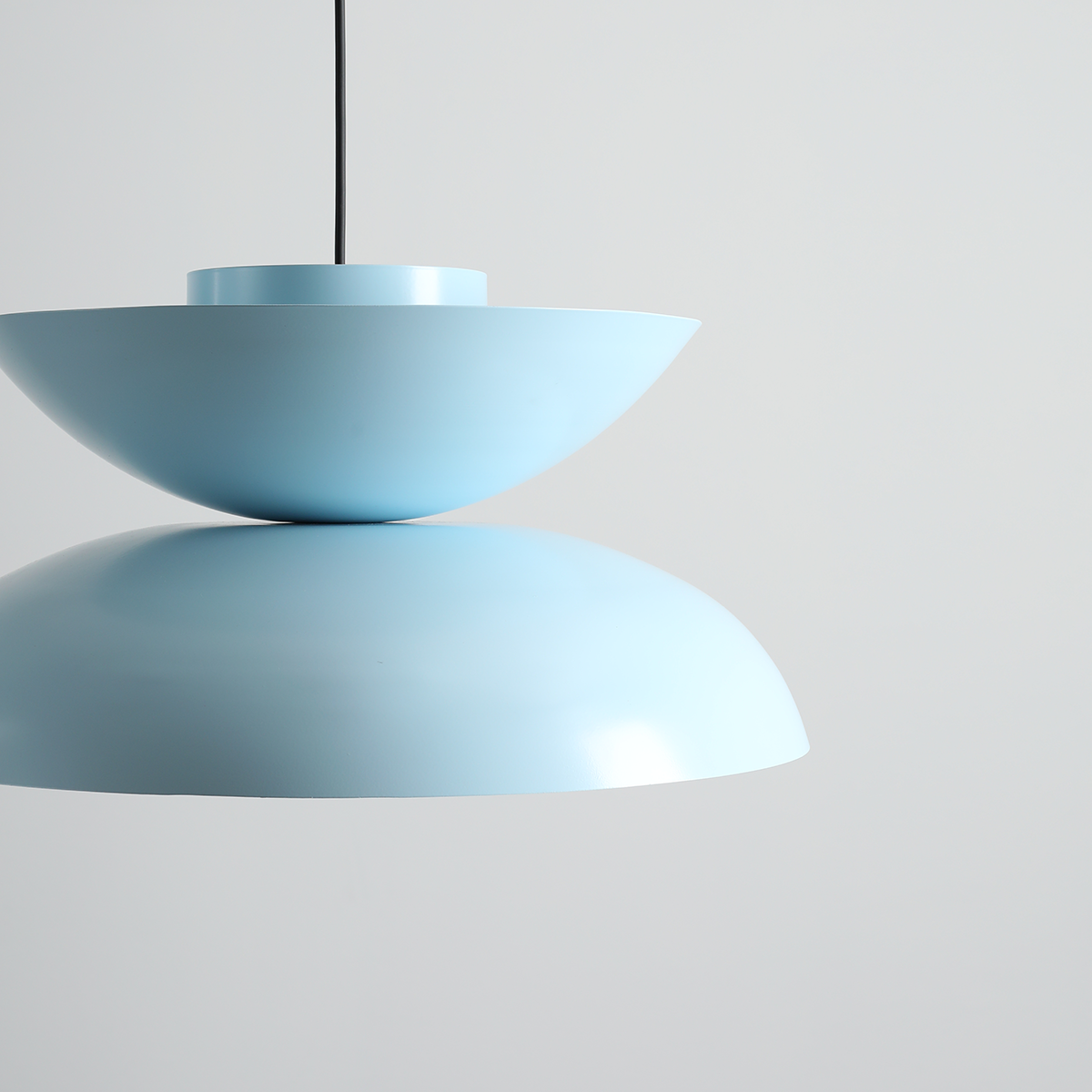 Lampa wisząca Millo w kolorze błękitu baby blue, złożona z dwóch półkulistych elementów. Górna część jest wklęsła, a dolna ma gładką, zaokrągloną formę. Zwisa na czarnym przewodzie, umieszczona na jasnym tle.