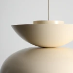 Lampa wisząca Millo w kolorze jasnej kremowej, o opływowym kształcie. Ujęcie z góry, pokazujące szeroką, płaską czaszę oraz centralny element mocujący. Lampa zawieszona na cienkim przewodzie, umieszczona na gładkim, lustrzanym tle.