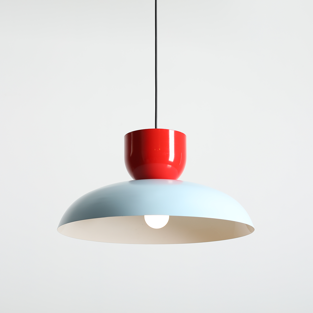 Lampa wisząca Ella z kloszem w kolorze błękitnym i czerwonym uchwytem. Wisi na czarnym przewodzie, z widoczną żarówką E27. Kształt okrągły, z matowym wykończeniem. Tło proste, jasne.