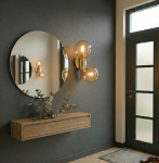 Kinkiet Eral 2xE14 w złotym, błyszczącym wykończeniu, z dwoma szklanymi kloszami, zamontowany na ciemnej ścianie obok okrągłego lustra. W tle widoczna drewniana półka z rośliną oraz drzwi z przeszklonymi panelami.