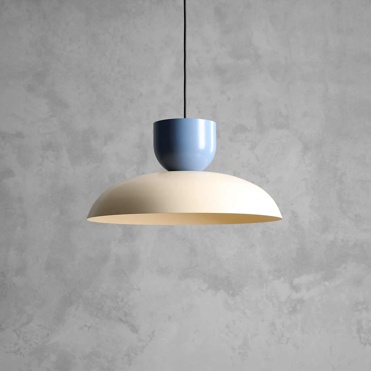 Lampa wisząca Ella z jednym gniazdem E27, z okrągłym kloszem w kolorze ciemnej kremowej i cylindrycznym elementem w odcieniu przybrudzonego niebieskiego. Zawieszona na czarnym przewodzie, na tle szarej ściany.