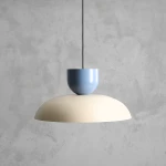 Lampa wisząca Ella z kloszem w kolorze ciemnej kremowej i niebieską podstawą. Ujęcie frontalne, produkt zawieszony na czarnym przewodzie. Gładka, zaokrąglona forma, idealna do nowoczesnych wnętrz. Tło w odcieniu szarości.