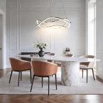 Lampa wisząca LED 36W CCT w kolorze złotym, o nowoczesnym, falistym kształcie, zawieszona nad okrągłym stołem w eleganckim wnętrzu. W tle widoczne są krzesła z tapicerowanymi siedzeniami w kolorze brązowym i dekoracyjny wazon z kwiatami.
