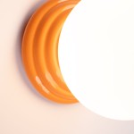 Kinkiet Zori 1xG9 IP44 w kolorze pomarańczowym, z okrągłą, białą kloszem. U podstawy widoczna gładka, błyszcząca podstawa z charakterystycznymi rowkami. Produkt prezentowany z bliska, na jasnym tle.