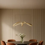 Lampa wisząca Snow 1xLED 40W CCT w złotym kolorze, o nowoczesnym, falistym kształcie. Zawieszona na cienkich linkach nad okrągłym stołem, w minimalistycznym wnętrzu z neutralnym tłem. Widać także szklany wazon z kwiatami.