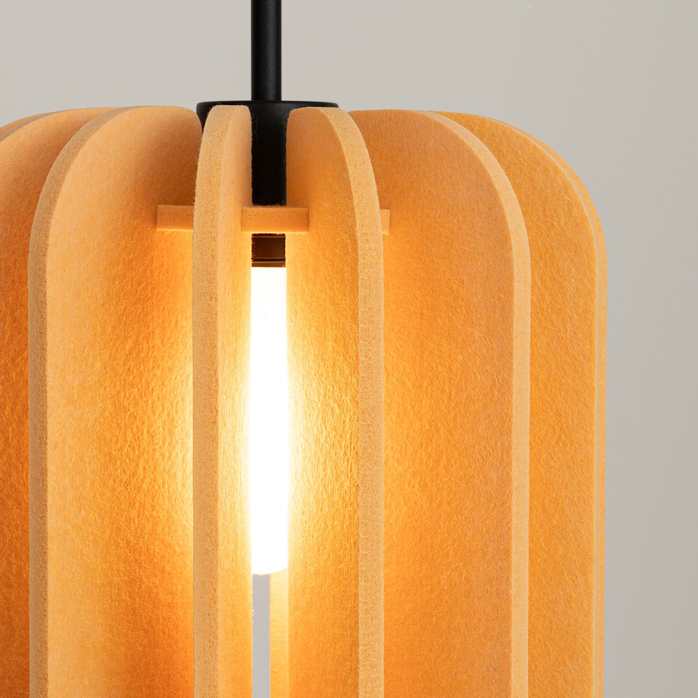 Lampa wisząca Mula D27 z czarnym stelażem i żółtym kloszem z materiału przypominającego filc. Klosz ma cylindryczny kształt z wycięciami, które tworzą efektowne oświetlenie. Widoczna żarówka E27 w środku.