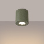 Lampa sufitowa Orbis Beton w kolorze zielonej oliwki, wykonana z betonu. Okrągły klosz z widocznym źródłem światła GU10, zamontowana na suficie, z fakturą przypominającą surowy beton. Tło w delikatnym, neutralnym odcieniu.