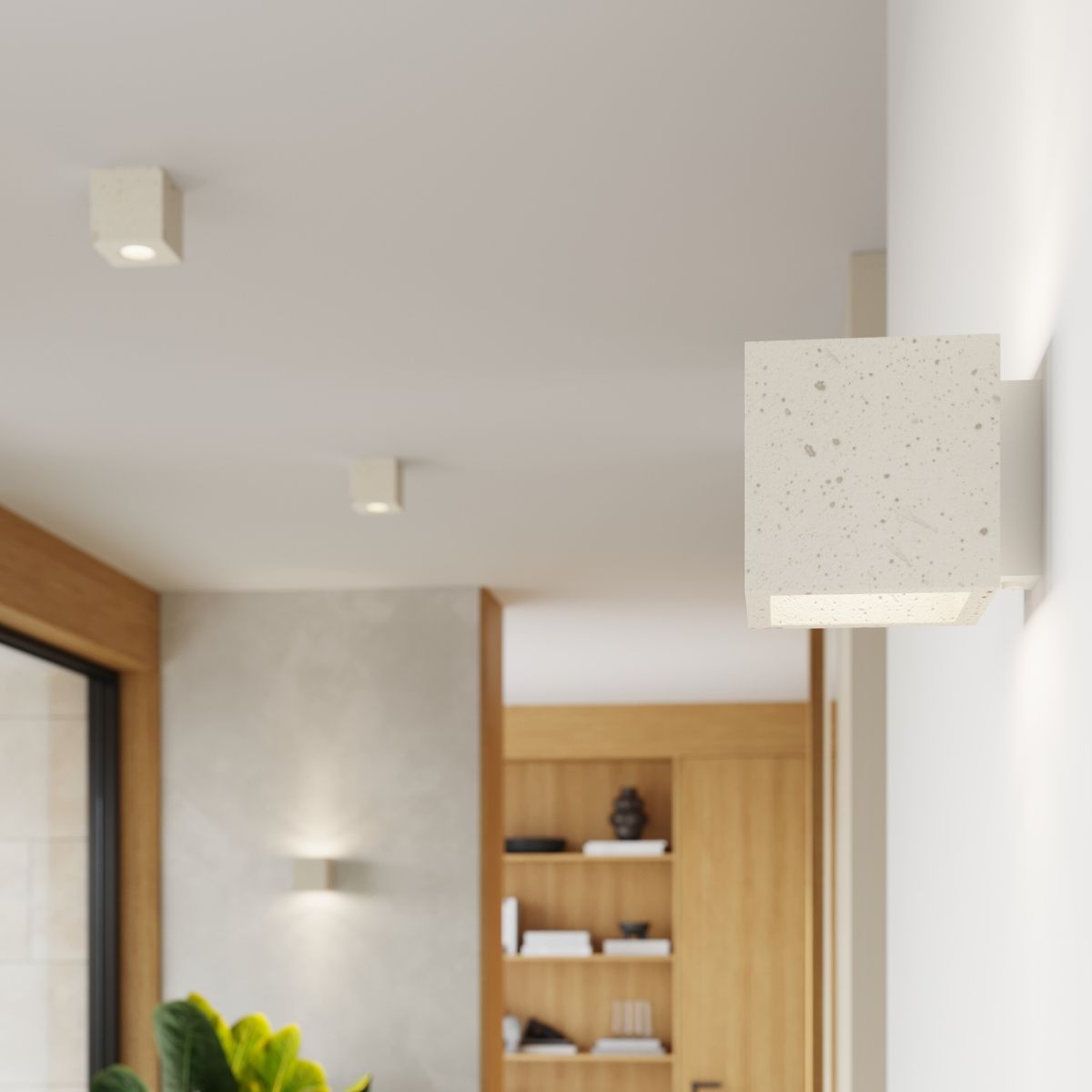 Kinkiet Quad Beton w kolorze beżowym, wykonany z betonu, o kwadratowej formie. Zamontowany na ścianie, z widocznym gniazdem na żarówkę G9. Tło w jasnych odcieniach, z drewnianymi akcentami oraz roślinnością w dolnej części kadru.