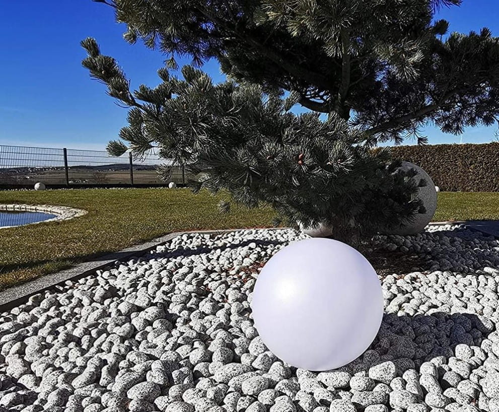 Ballo lampa solarna ogrodowa w kształcie dużej, białej kuli o średnicy 25 cm i wysokości 60 cm, umieszczona na żwirowym podłożu w ogrodzie, w otoczeniu drzew i zielonej trawy.