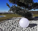 Ballo lampa solarna ogrodowa w kształcie dużej, białej kuli o średnicy 25 cm i wysokości 60 cm, umieszczona na żwirowym podłożu w ogrodzie, w otoczeniu drzew i zielonej trawy.