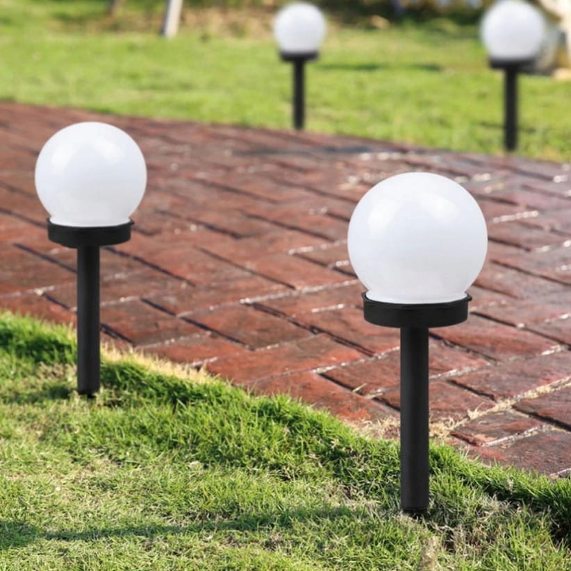 Lampa solarna ogrodowa Ballo D10/H34 z białą, okrągłą głowicą, umieszczona na czarnym stelażu. Widoczna na trawniku obok ścieżki z cegły. Wysokość lampy wynosi 34 cm, średnica głowicy 10 cm.