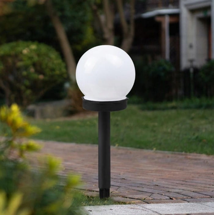 Ballo lampa solarna ogrodowa w kształcie białej kuli na czarnym słupku. Wysokość 34 cm, średnica 10 cm. Umieszczona na trawniku, w tle widoczna zieleń i kostka brukowa. Idealna do oświetlenia ogrodu.