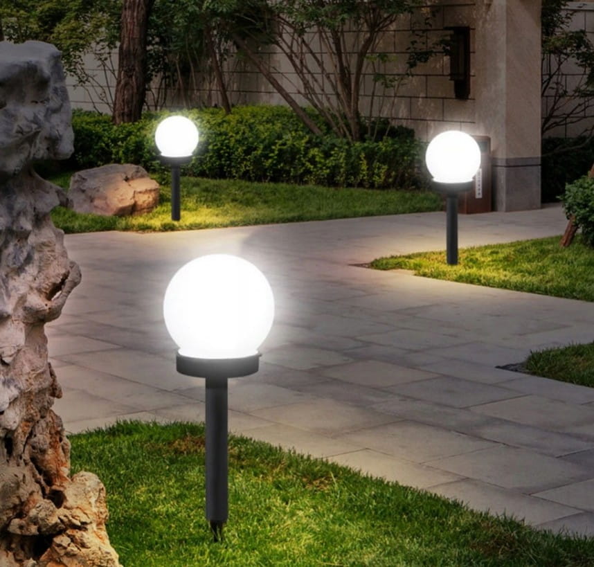 Ballo lampa solarna ogrodowa D10/H34 z białym, okrągłym kloszem, umieszczona na czarnym słupku. Widoczna w ogrodzie, na ścieżce, w otoczeniu trawy i kamieni. Lampy świecą ciepłym światłem, tworząc przyjemną atmosferę.