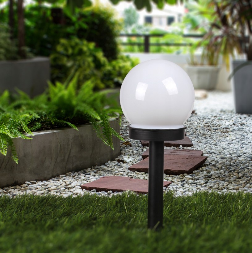 Lampa solarna ogrodowa Ballo D10/H34 z białym, okrągłym kloszem, umieszczona na czarnym słupku. Widoczna w ogrodzie, otoczona roślinnością i kamiennym podłożem. Klosz emitujący światło LED, stworzona do oświetlenia przestrzeni zewnętrznych.