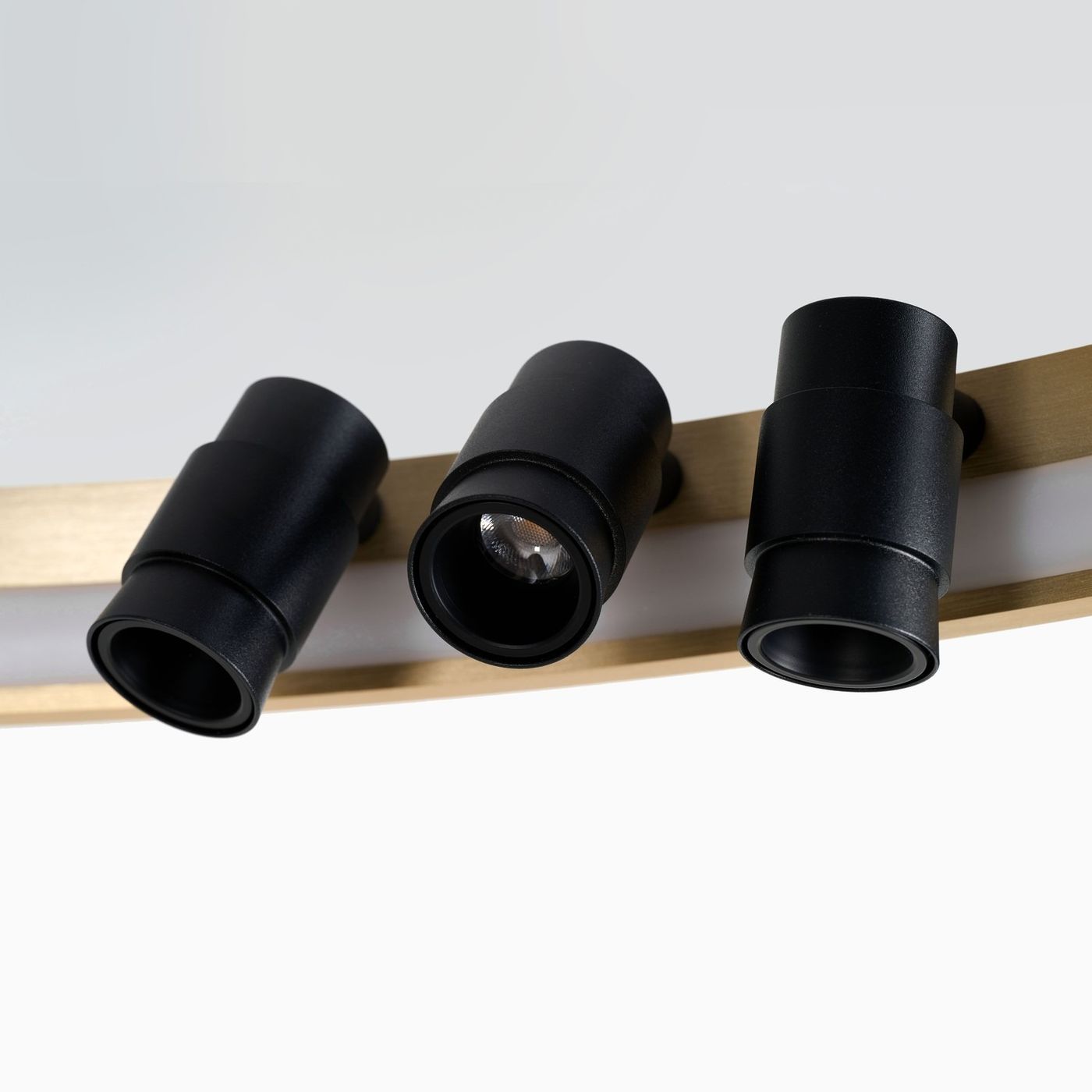 Lampa sufitowa Serafino D62 z siedmioma źródłami LED o mocy 34W. Czarna obudowa reflektorów na złotej podstawie. Widok z boku, ukazujący trzy czarne cylindryczne lampy skierowane w różne strony. Tło jasne, jednolite.