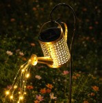 Konewka lampa solarna ogrodowa H80 LED, w kształcie konewki z metalową, ażurową obudową w kolorze złotym. Umieszczona na metalowym stelażu, emituje ciepłe światło LED, z dekorem imitującym wodę wypływającą z dzióbka. Tło z kwiatami.