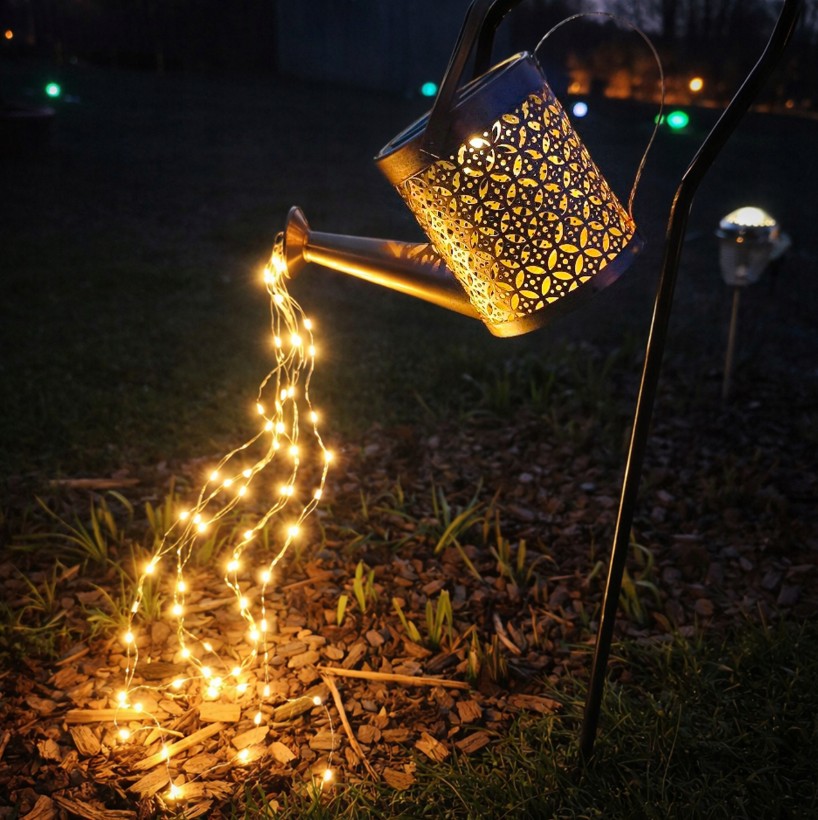 Konewka solarna ogrodowa z lampą LED, wbijana, w kształcie konewki z metalowym, ażurowym wzorem. Wydobywające się z niej ciepłe światło LED tworzy efekt wylewania na podłożu z koralików. Produkt umieszczony na stelażu, w ogrodzie, po zmroku.