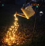 Konewka solarna ogrodowa z lampą LED, wbijana, w kształcie konewki z metalowym, ażurowym wzorem. Wydobywające się z niej ciepłe światło LED tworzy efekt wylewania na podłożu z koralików. Produkt umieszczony na stelażu, w ogrodzie, po zmroku.