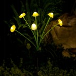 Grzybki lampa solarna ogrodowa, wbijana, z pięcioma żółtymi kloszami LED na długich, zielonych nóżkach. Ustawiona w nocy w ogrodzie, otoczona roślinnością, emituje ciepłe światło, tworząc przytulną atmosferę.