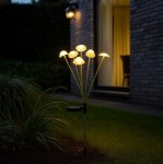 Lampa solarna ogrodowa w formie grzybków, z sześcioma delikatnie świecącymi kloszami w ciepłym odcieniu żółtego. Ustawiona w pionie, z metalowym stelażem i podstawą do wbicia w ziemię. Tło z ciemnym murem i oświetlonym wnętrzem budynku.