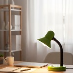 Zielona lampka biurkowa Mentor z regulowanym ramieniem i podstawą, wyposażona w oprawę E27. Ujęcie z boku, na biurku, obok filiżanki z parującym napojem oraz notatnikiem. Tło jasne, z zasłonami i półką w tle.