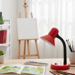 Czerwona lampka biurkowa Mentor z elastycznym ramieniem, 1xE27. Ustawiona na drewnianym biurku obok otwartej książki z ilustracjami, zestawu farb oraz pędzli, w tle widoczna sztaluga i okno z zasłonami.