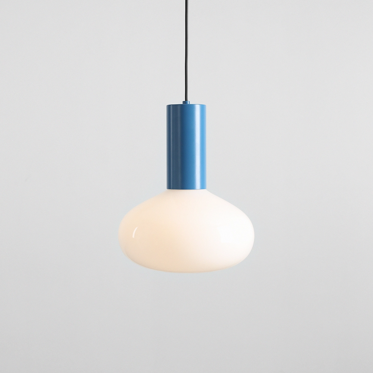 Lampa wisząca Flo Sky D26 z niebieską metalową podstawą i białym szklanym kloszem w kształcie kulistym. Zawieszona na cienkim czarnym przewodzie, prezentuje nowoczesny design. Tło w jasnym kolorze, proste i minimalistyczne.