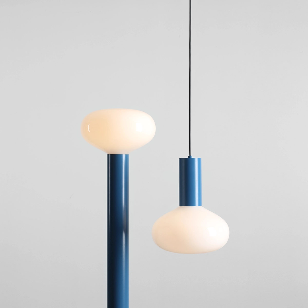Lampa wisząca Flo Sky D26 w niebieskiej obudowie z metalowym wykończeniem, z mlecznym kloszem w kształcie kulistym. Produkt zawiera jedno gniazdo E14 i jest zawieszony na cienkim czarnym kablu, na jednolitym jasnym tle.