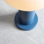 Mała lampa stołowa Flo Sky z niebieską, matową podstawą i okrągłym kształtem. Górna część lampy jest w kolorze jasnym, tworząc kontrast z podstawą. Ujęcie z góry, na tle jasnego, gładkiego materiału.